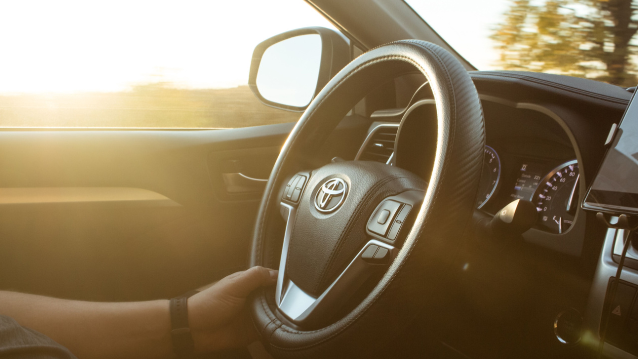 Man is driving vehicle with sun flare from the outside in the sunny day. Toyota sign on car steering wheel.