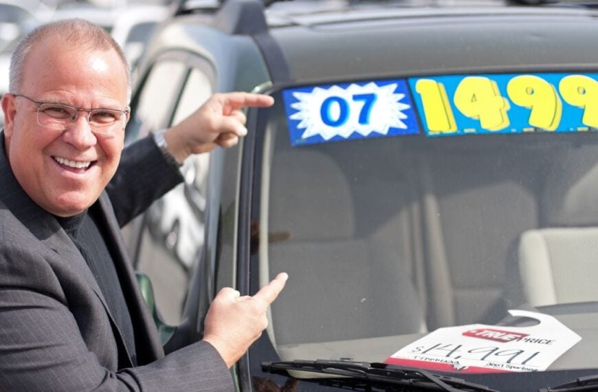 A middle-aged white male pointing to the sale price of a used car.
