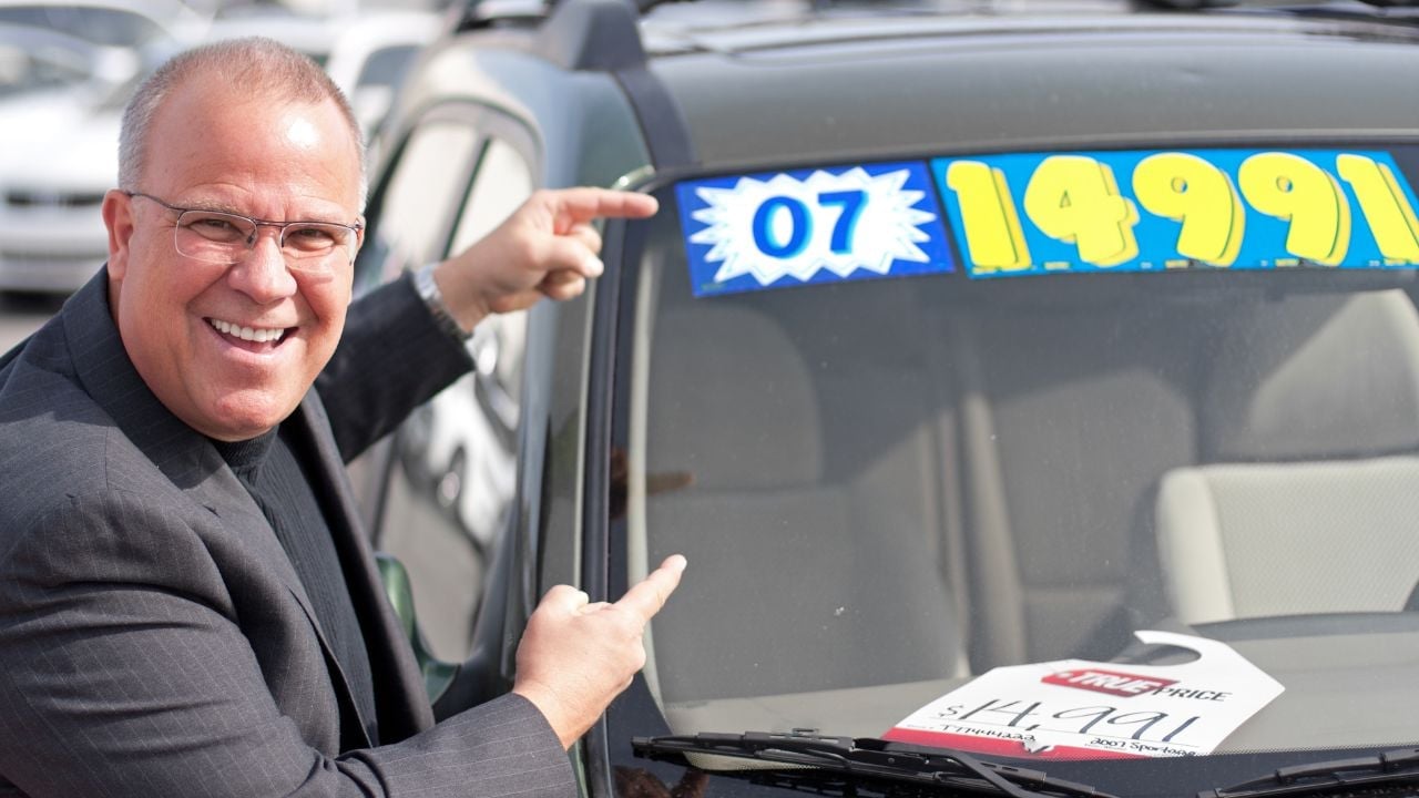 A middle-aged white male pointing to the sale price of a used car.