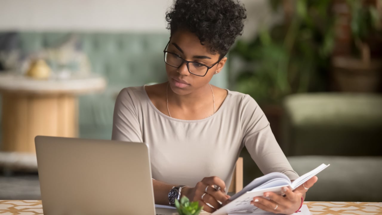 Focused young african american businesswoman or student looking at laptop holding book learning, serious black woman working or studying with computer doing research or preparing for exam online