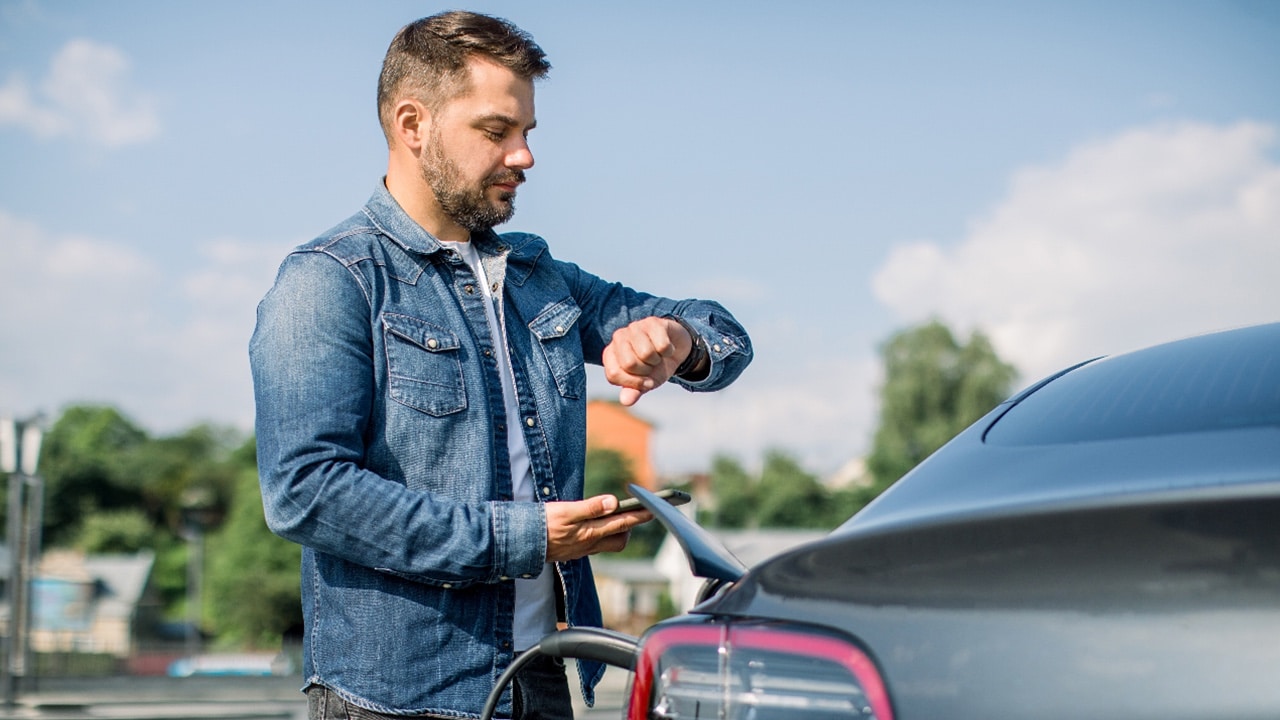 Young Caucasian bearded man standing near the electric car and looks at the watch and smart phone. His electric car is charging at the city charging station for electric vehicles.