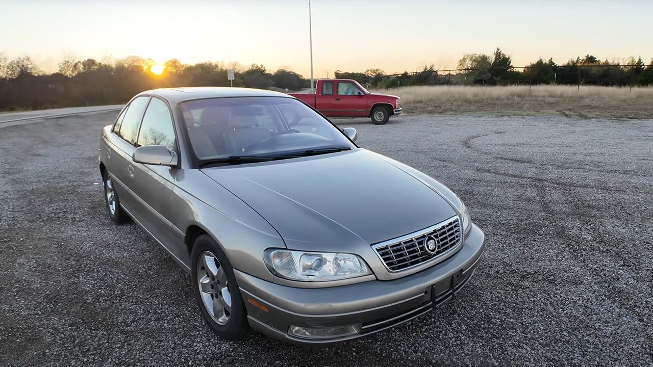 A light brown Cadillac Catera in front of a sunset, front 3/4 view