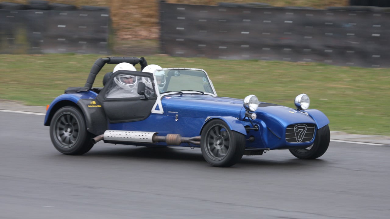 A blue Caterham Seven with the optional canvas doors on a track day, front 3/4 view