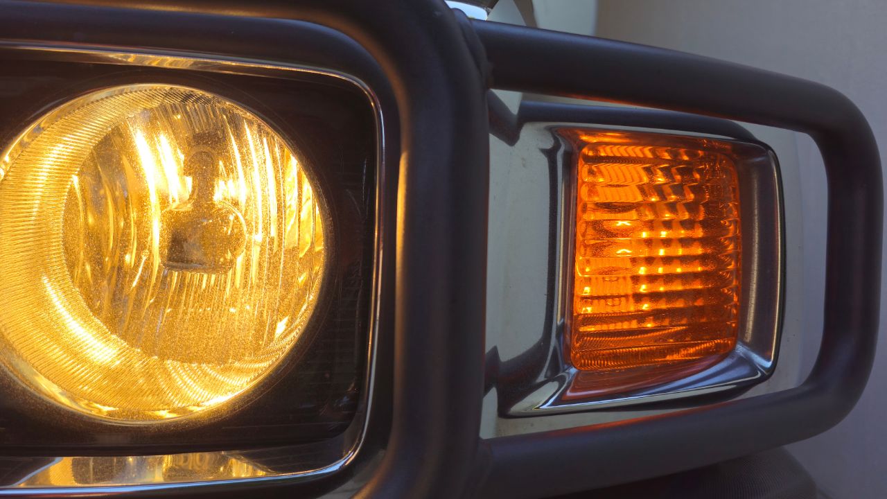 A close-up of a classic off-road vehicle's glowing headlight and turn signal, featuring a rugged metal grille. The warm amber and yellow lights create a nostalgic, adventure-ready atmosphere.