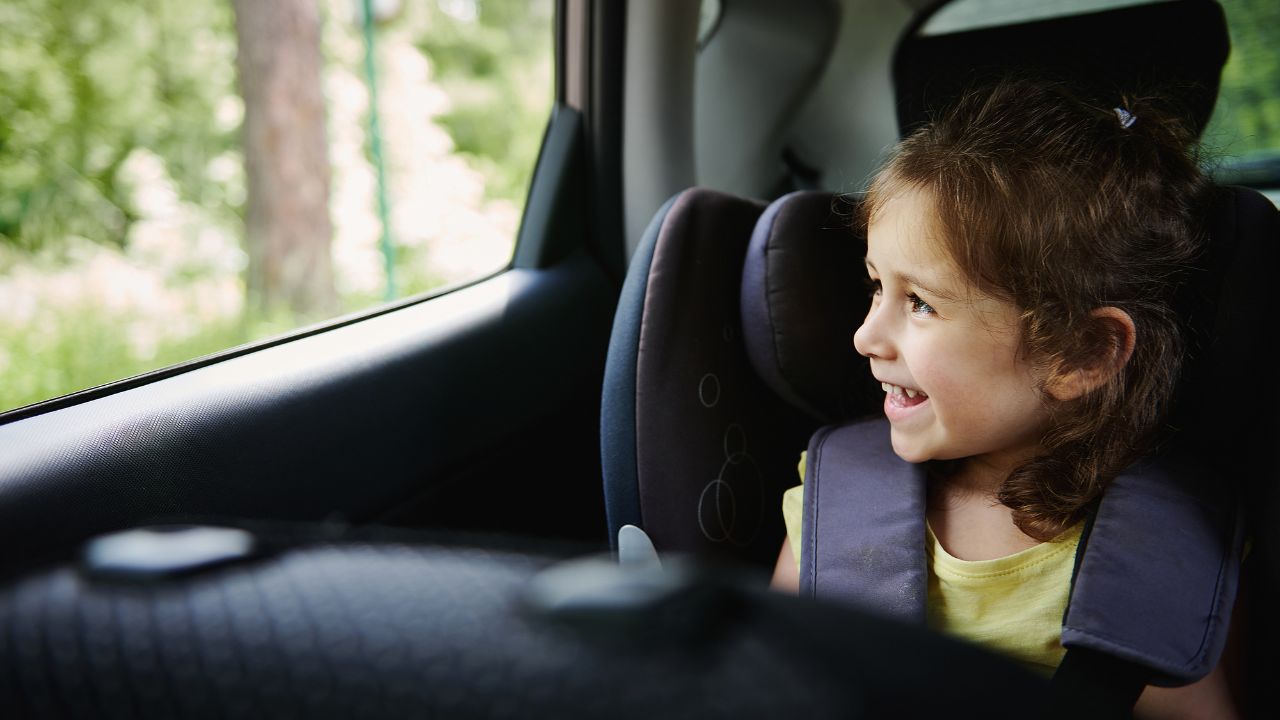 Safe movement of children in the car. Adorable baby girl enjoys the travel by car, looks out of an opened window while sitting in a safety booster car seat