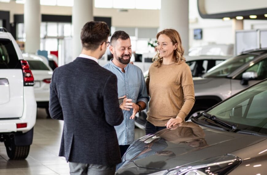 Young car salesman showing to mid-age couple new automobile at dealership salon