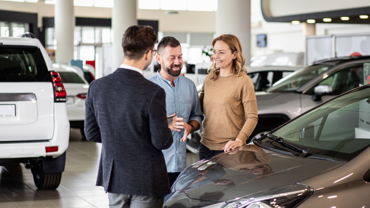 Young car salesman showing to mid-age couple new automobile at dealership salon