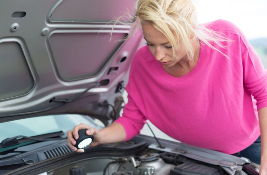 Self-sufficient confident modern young woman inspecting broken car engine with a flashlight in her hand