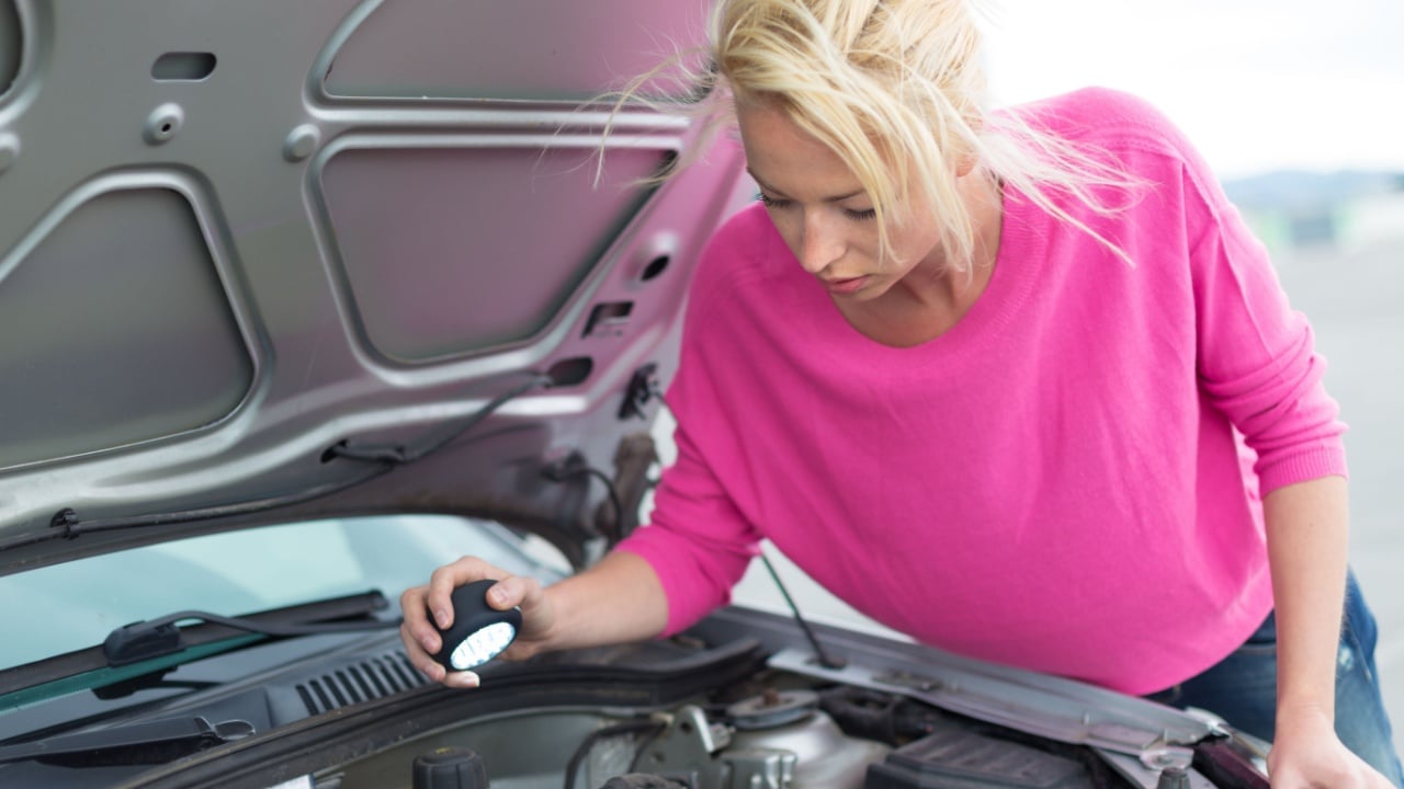 Self-sufficient confident modern young woman inspecting broken car engine with a flashlight in her hand
