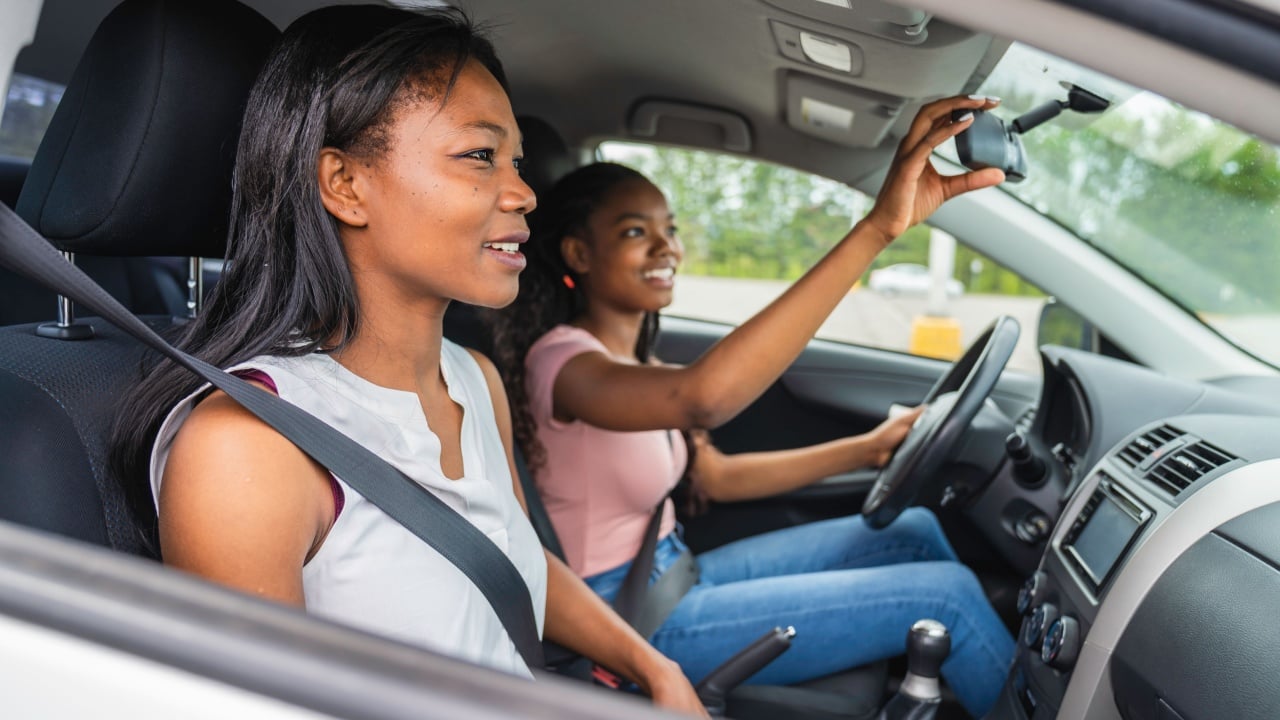 Teenage driver adjusts the mirror of her car. Her mother is in the passenger seat
