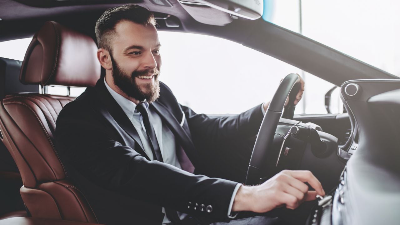 Young handsome businessman is sitting in luxury car. Serious bearded man in suit is driving.
