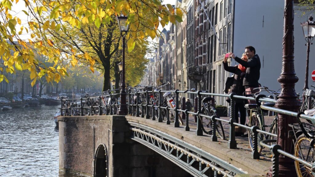 Couple take a selfie on a bridge while a tour boat sails under the bridge over a canal with autumn trees in historical Amsterdam, The Netherlands