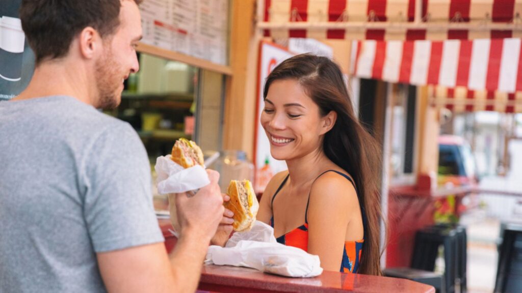 Happy couple eating sandwiches in typical retro cafe in Florida. Cuba sandwich local food. Summer travel tourist lifestyle young Asian woman smiling eating lunch outside