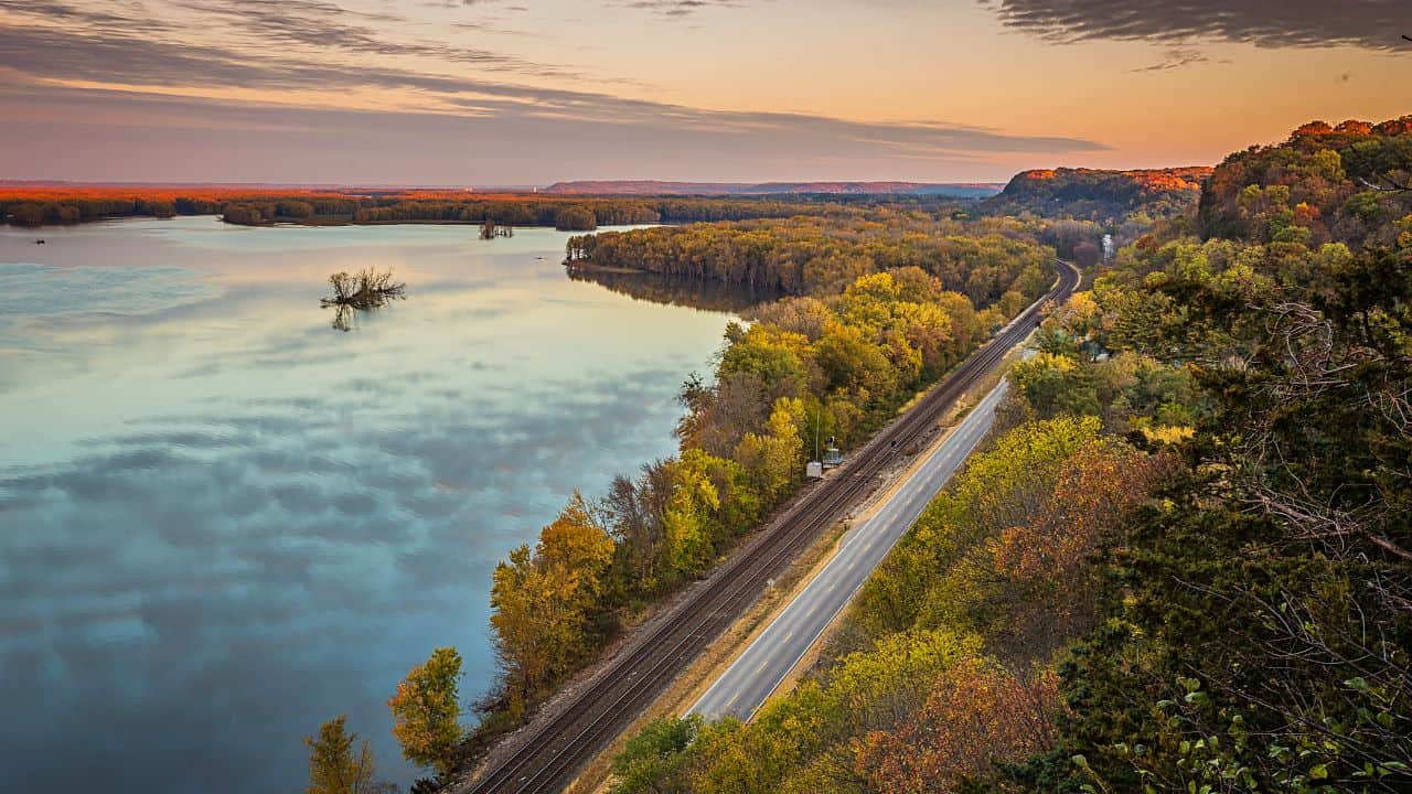 Scenic river view at sunset with a railroad track alongside, surrounded by lush autumn foliage.