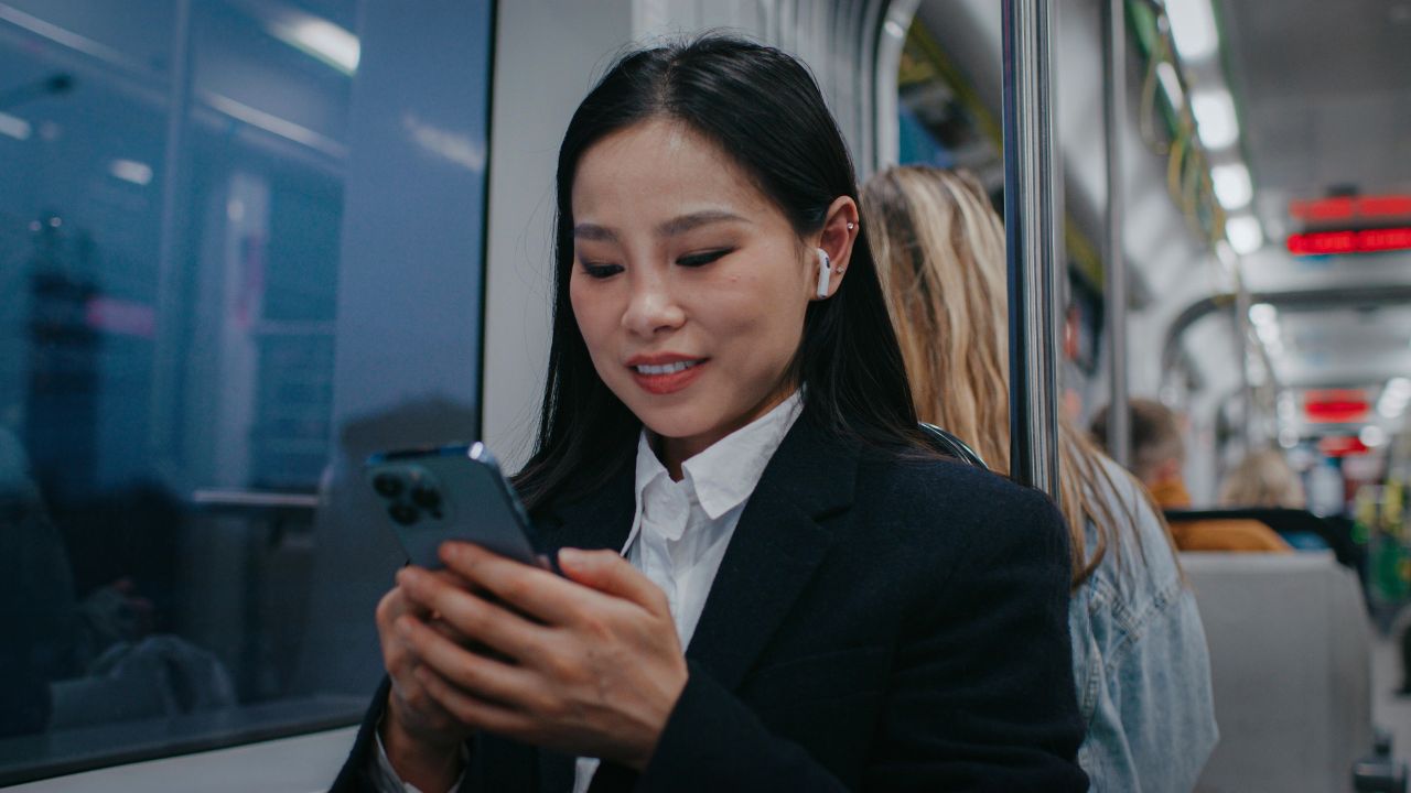Positive Asian female wearing earphones while holding her smartphone. Pretty girl having chat with someone on phone. Sitting near window during trip on tram or bus. Remote communication.