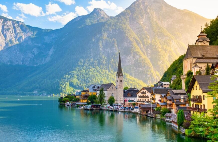 Amidst towering mountains, charming houses imbue Hallstatt Austria with vibrant colors along the lakeside. The golden sunlight dances on calm waters, creating a picturesque reflection at dusk.