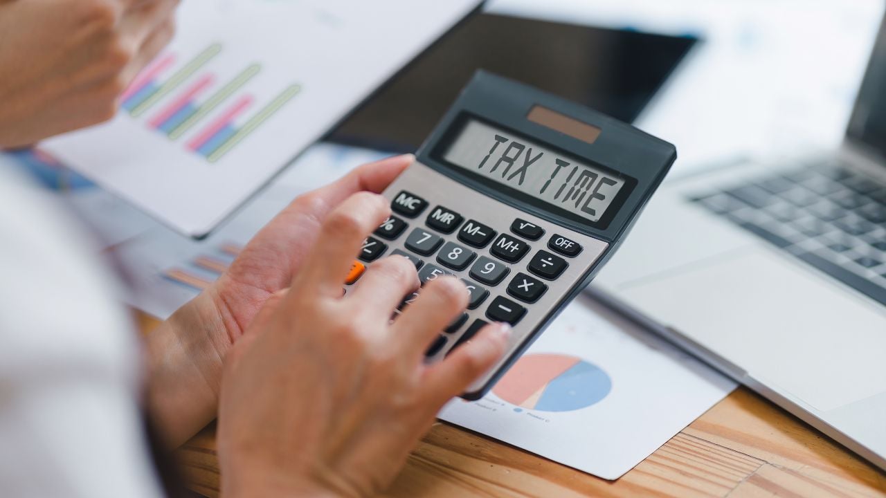 Close-up of hands using calculator with "TAX TIME" on display, alongside financial charts and a laptop. Calculating Finances During Tax Season
