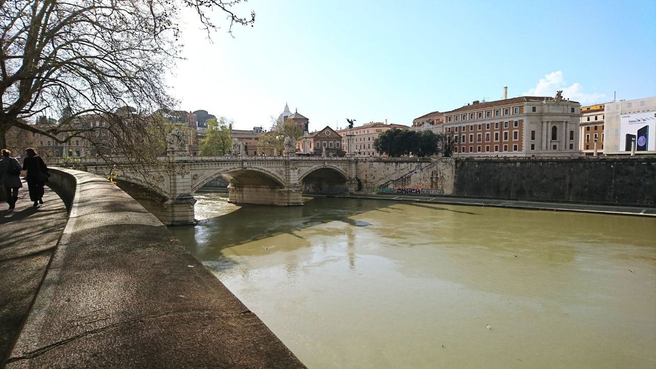 Vatican City - April 05 2018: People walking along the promenade of the Tiber River by the historic Ponte Vittorio Emanuele II bridge with the city's famous dome in the distance on a clear day