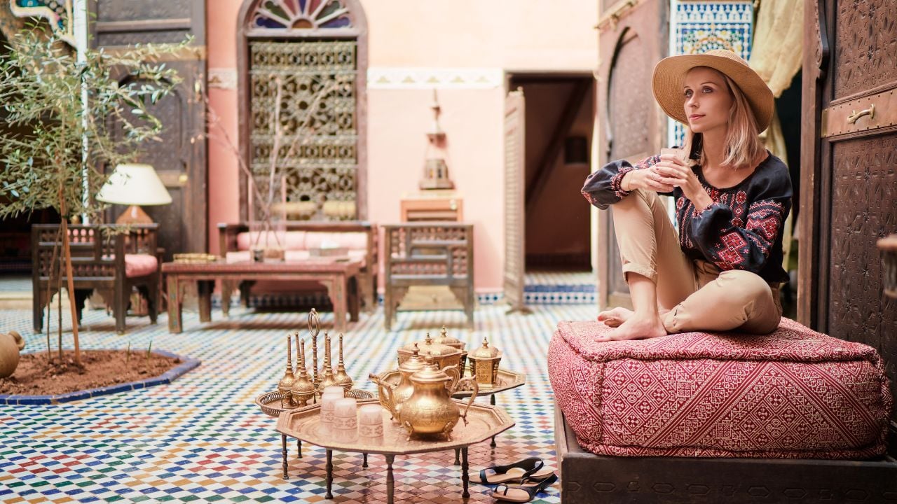 Traveling by Morocco. Happy young woman in hat relaxing in traditional riad interior in medina.