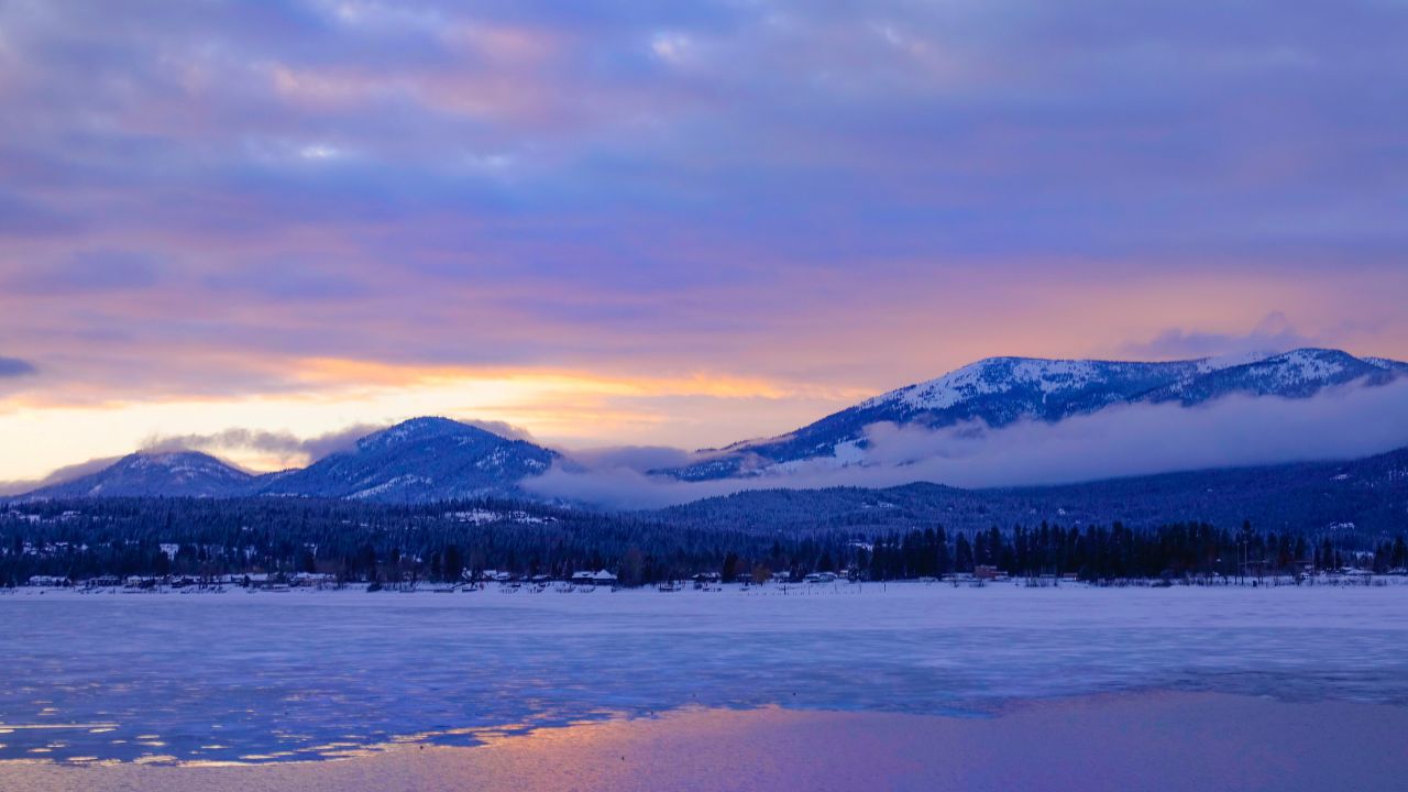 Picturesque view of Lake Pend Oreille and adjacent mountains at golden sunrise. Clouds gather over a large frozen lake under a small forest covered ridge in Idaho, USA. Winter scenery on sunny morning