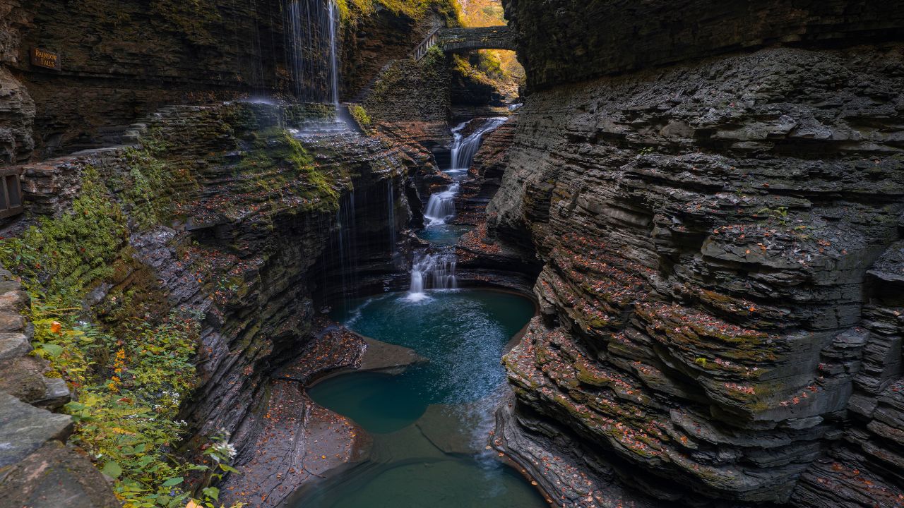 Stone bridge over river and cascades in deep limestone gorge, hiking trail going along, fallen colorful fall leafs surrounding the creek. Wide angle, long exposure. Watkins Glen State Park, NY, USA.