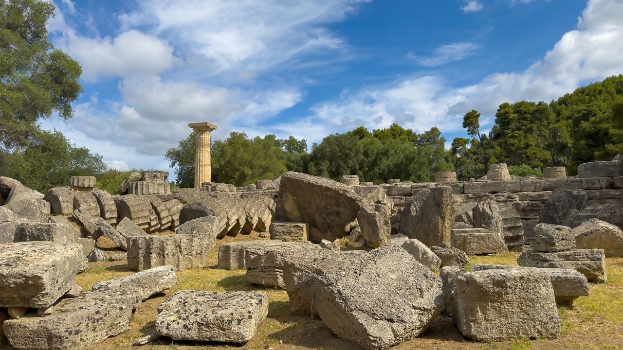 Olympia, Greece - July 4, 2024: Birthplace of the Olympic Games. Heap of column pieces in front of Temple of Zeus with the lone standing column visible, Green foliage and thick blue cloudscape