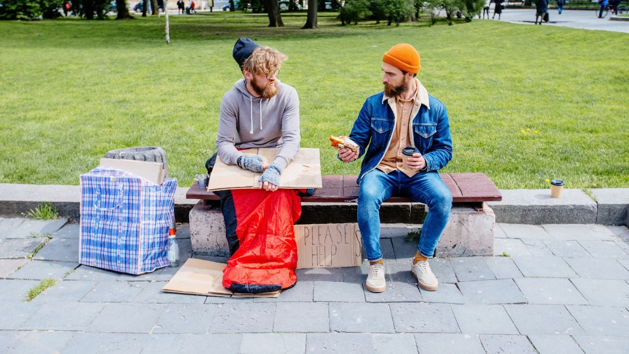 Opposite concept. Handsome beard man sits on bench with coffee and croussant proposing food for strange hungry homeless man in sleeping bag near him. Concept of helping poor people