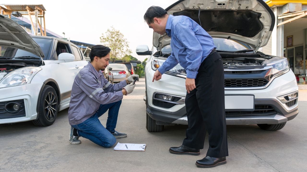 Two individual inspect damage on a vehicle. Car’s owner pointing at a dent on the bumper of the car. An insurance staff taking picture with smartphone, conducting an assessment related to the damage.