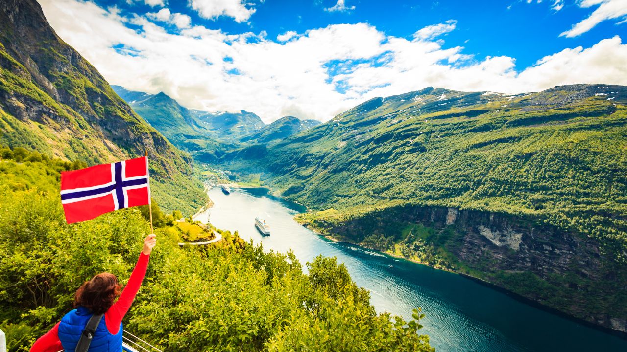 Female tourist with norwegian flag enjoying scenic view over fjord Geirangerfjord with cruise ship. Cruising vacation and travel.
