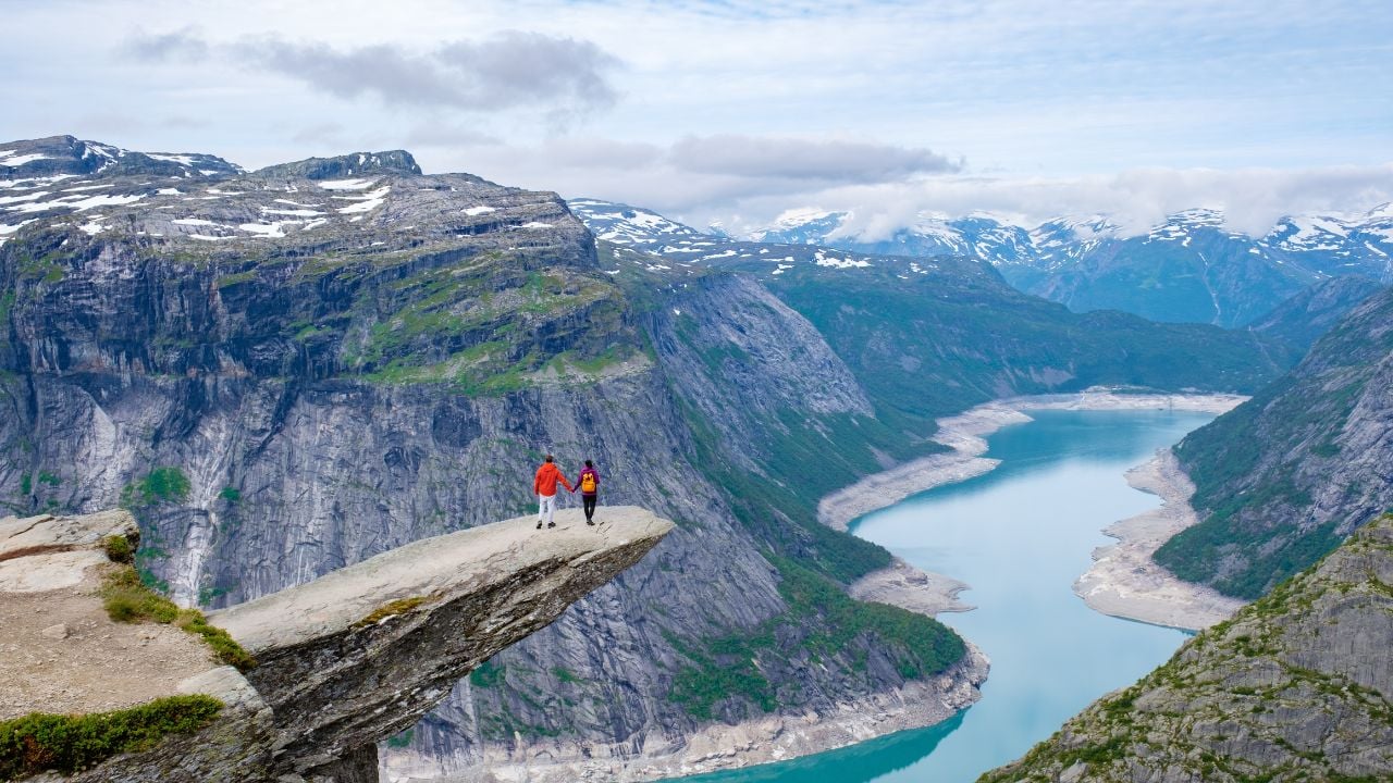 A couple stands on the edge of Trolltunga, Norway, a famous cliff in Norway, overlooking a breathtaking view of the surrounding mountains and a winding lake.