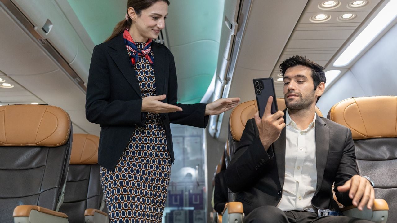 Flight attendant interacts with male passenger talking on mobile phone during boarding or pre-flight procedure inside commercial airplane cabin.