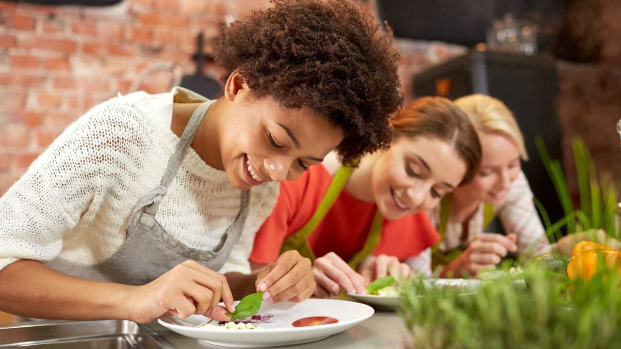 cooking class, friendship, food and people concept - happy women cooking and decorating plates with dishes in kitchen