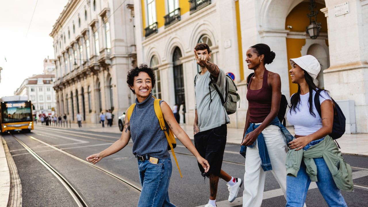 Four happy tourists with backpacks are walking and crossing the street in lisbon near a tram, a man is pointing his finger showing something to his friends