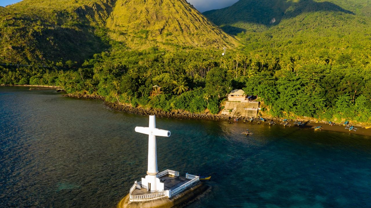 Aerial view of a large memorial cross marking a sunken cemetery in golden evening sunlight (Camiguin, Philippines)