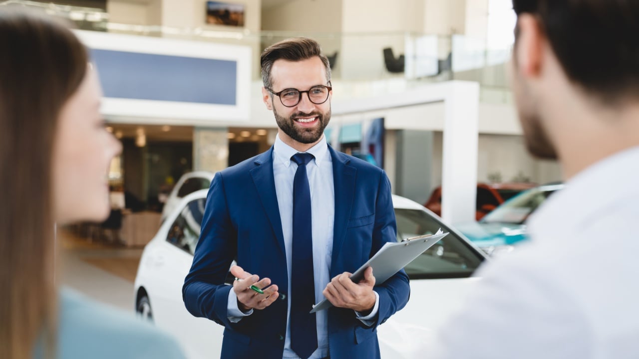 Focused photo of young male shop assistant explaining talking offering buying new car to young caucasian family husband and wife at auto dealer shop.