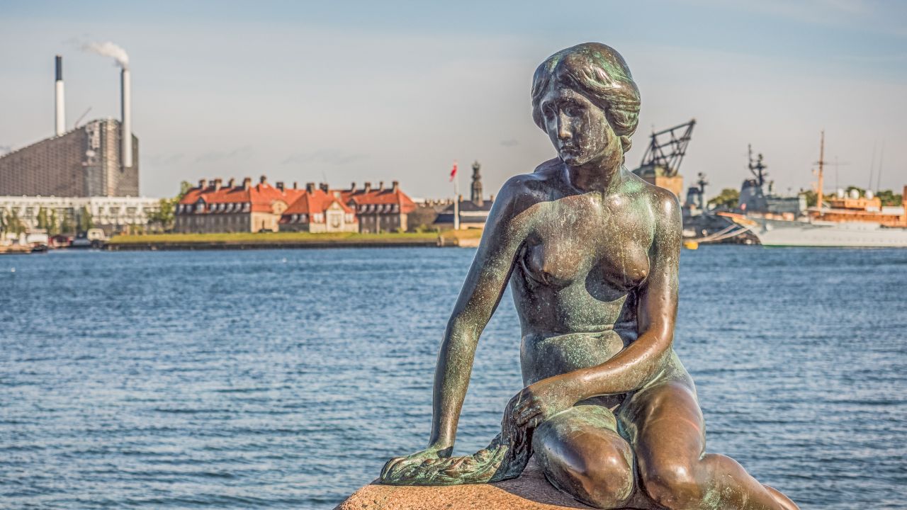 Copenhagen, Denmark - May 31, 2018: Bronze statue of the Little Mermaid, symbol of Kopenhagen, Den lille Havfrue, on the coastal rocks, with nuclear power plant in the background