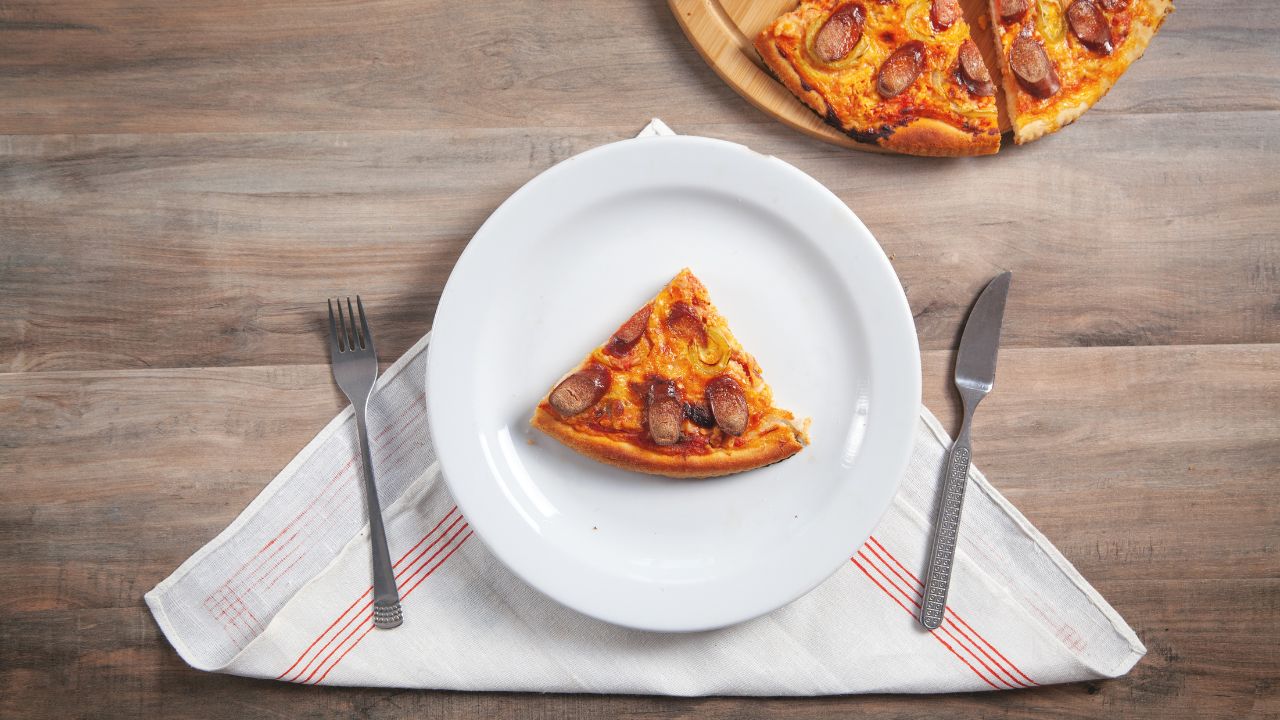 Slice of pizza on plate, fork, knife on wooden table. Pizza on circle board