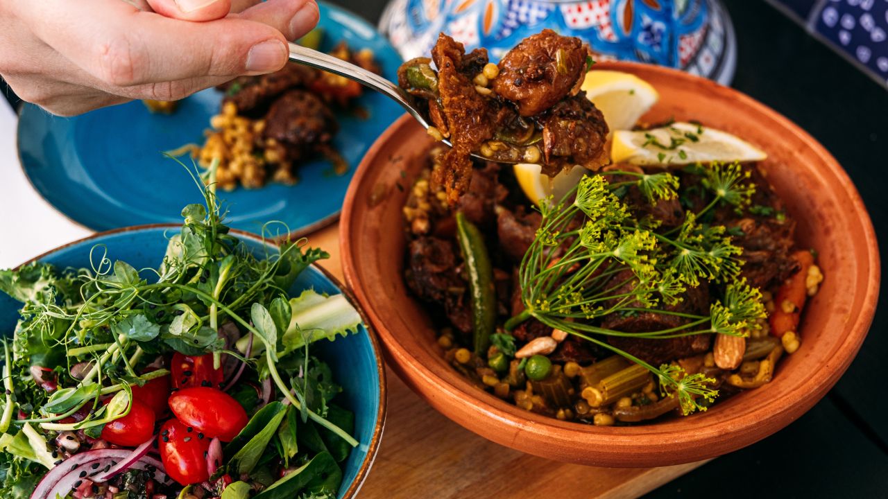 Traditional Moroccan lamb tagine served with a fresh salad. A hand scoops the dish, highlighting Mediterranean flavors and cultural culinary practices.