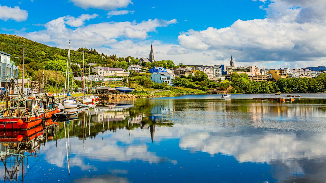 Pier at the port of Clifden at high tide, boats anchored with mirror reflection in the water, townscape against sky, sunny spring day with a blue sky and abundant white clouds in Clifden, Ireland