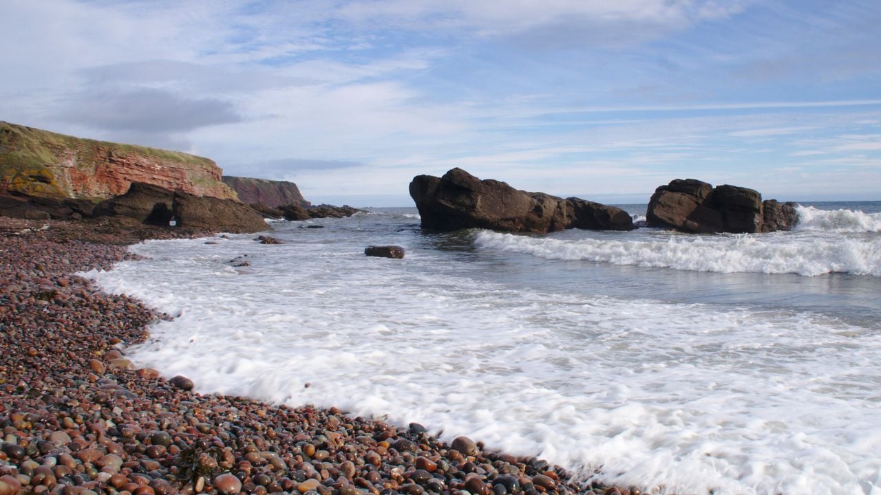 Waves washing up a pebble beach, with rocks and a cliff in the background. Auchmithie Harbour, Scotland.