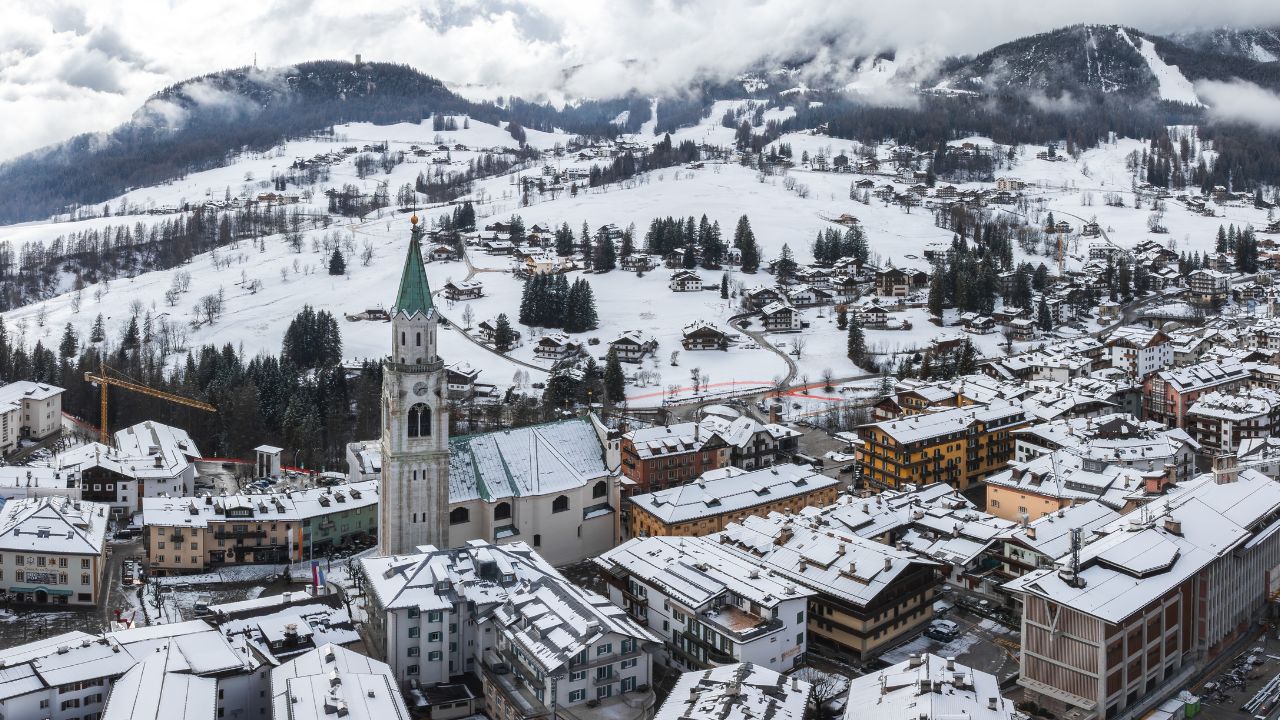 Aerial view of Cortina d'Ampezzo, Italy, featuring the Basilica Minore dei Santi Filippo e Giacomo, snow covered rooftops, and misty Dolomite peaks.