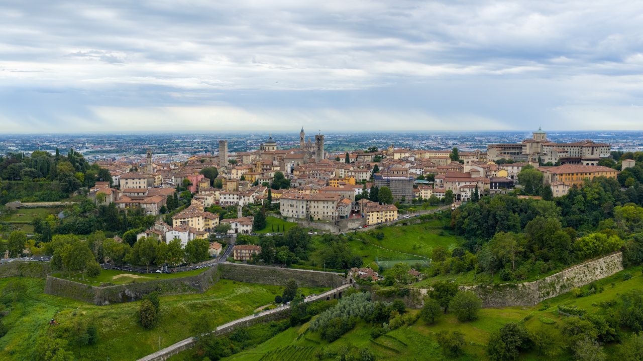 Drone panorama of Bergamo’s Città Alta hilltop skyline above terraced greenery under overcast summer clouds in Lombardy.