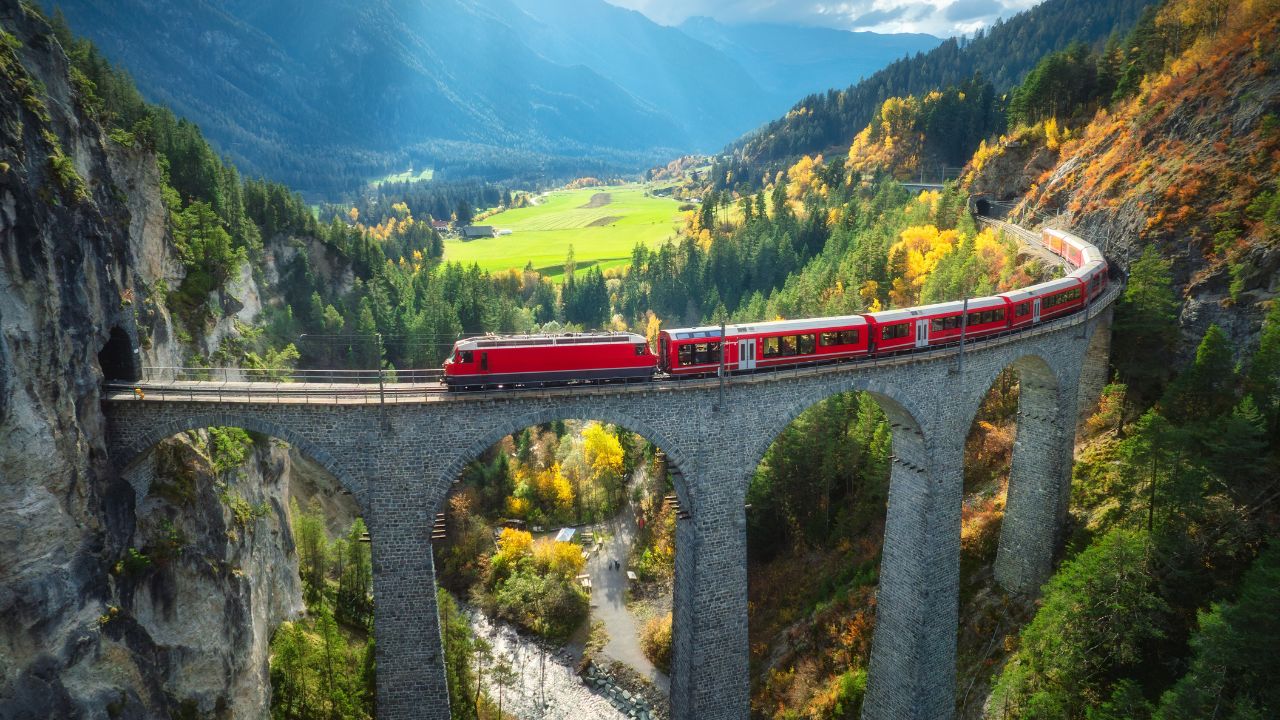 Aerial view of modern red train on Landwasser viaduct in alpine mountains, colorful forest at sunset in autumn. Bernina Express, Switzerland. Top view of train in Alps, railroad, green trees in fall