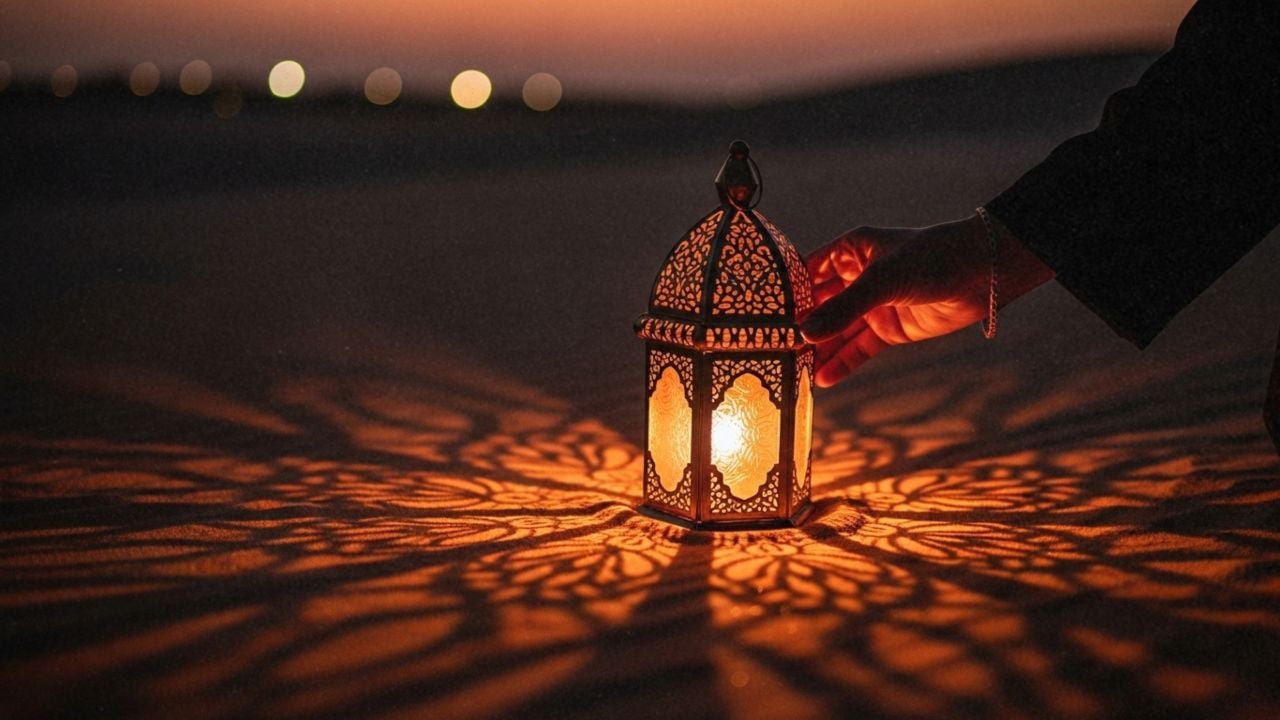 An Emirati woman's hand places a glowing traditional Ramadan lantern on a sandy surface. The low-light, warm-toned image with cinematic shadows evokes a mood of culture, spirituality, and mystery