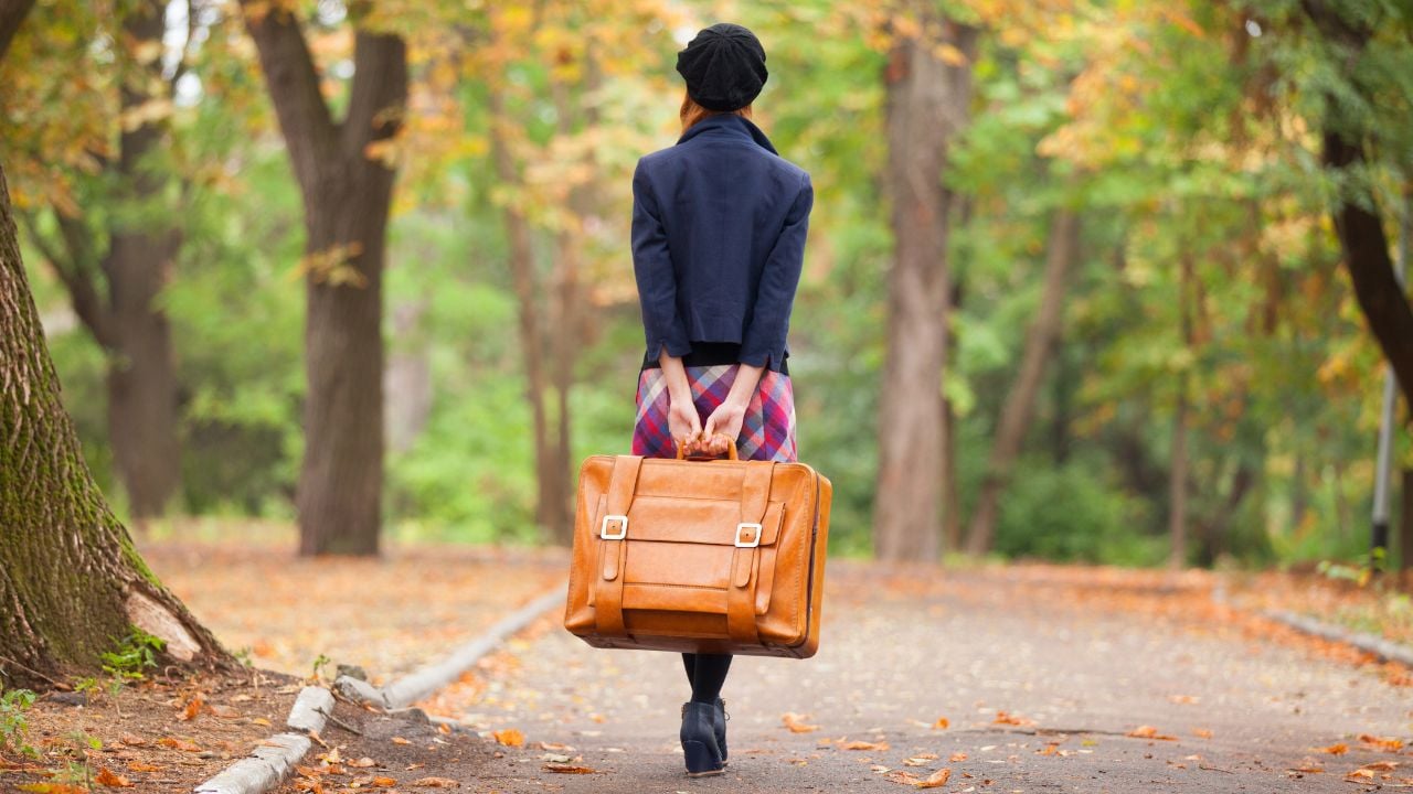 Woman dressed in coat (clothes) walking with orange suitcase in autumn in the park