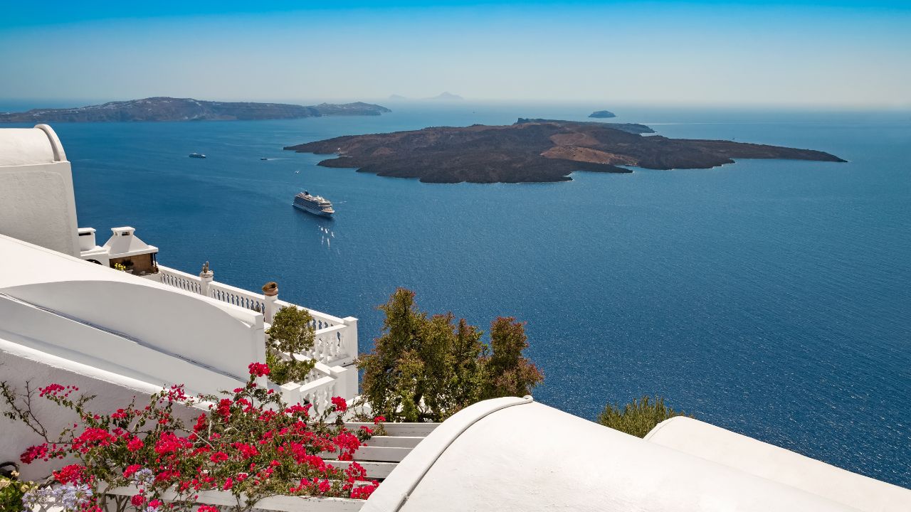 The caldera of Santorini, Greece, showcasing deep blue waters of the Mediterranean, the volcanic Nea Kameni island and a cruise ship, framed by Santorini's iconic white architecture and vibrant bloom.