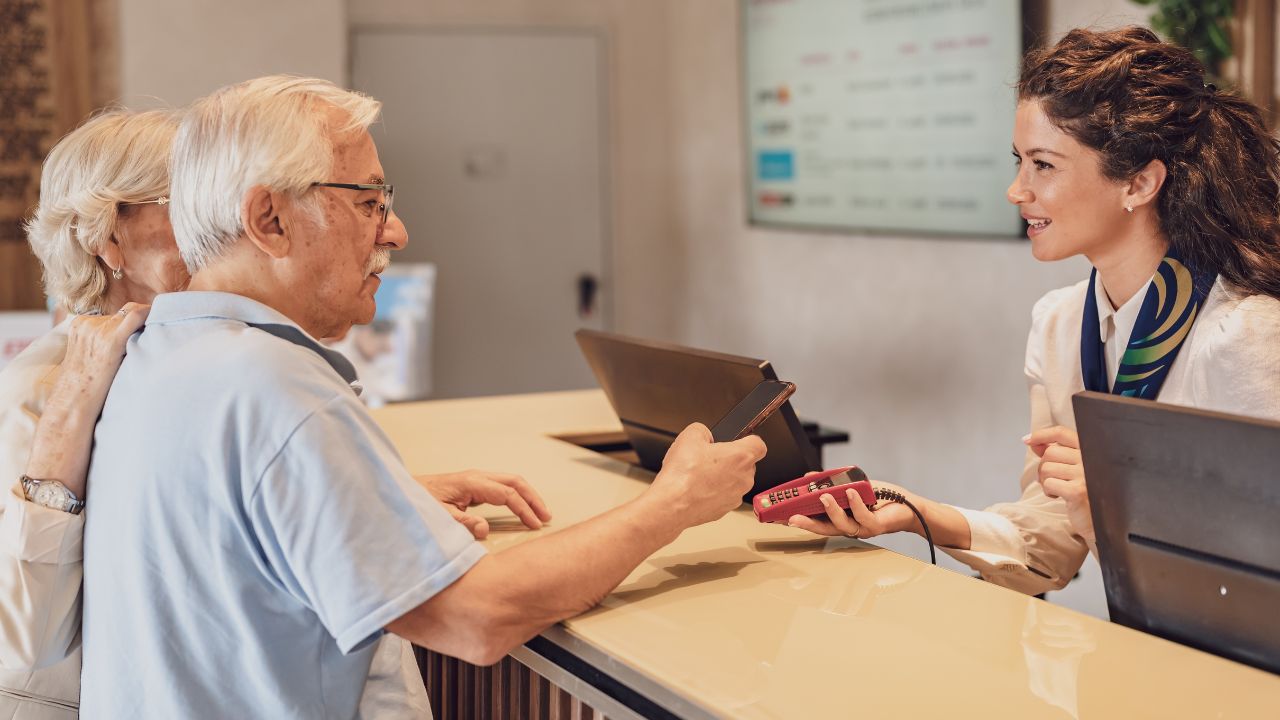 A senior couple at the hotel front desk makes contactless payments with a smartphone. Seniors traveling and using new technologies