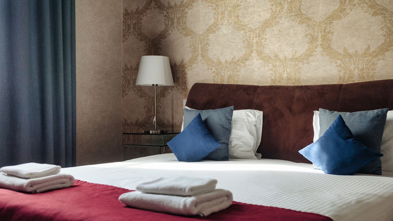 A comfortable Queen bed is staged in a Hotel room, layered with pillows, blankets, and towels. A bedside table and lamp stand next to a wall covered in an ornate Wallpaper pattern.