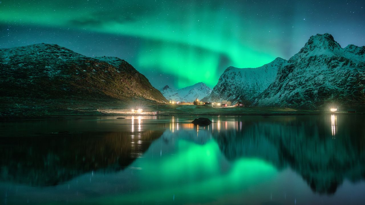 Aurora borealis, snowy mountains, sea, fjord, reflection in water, street lights at starry winter night. Lofoten, Norway. Northern lights. Landscape with polar lights, snowy rocks, sky with stars