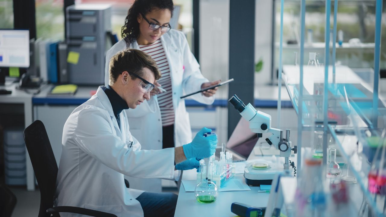 Modern Medical Research Laboratory: Portrait of Two Young Scientists Mixing Liquid Chemicals Using Pipette, Digital Tablet, Talking. Diverse Team of Multi-Ethnic Specialists work in Advanced Lab